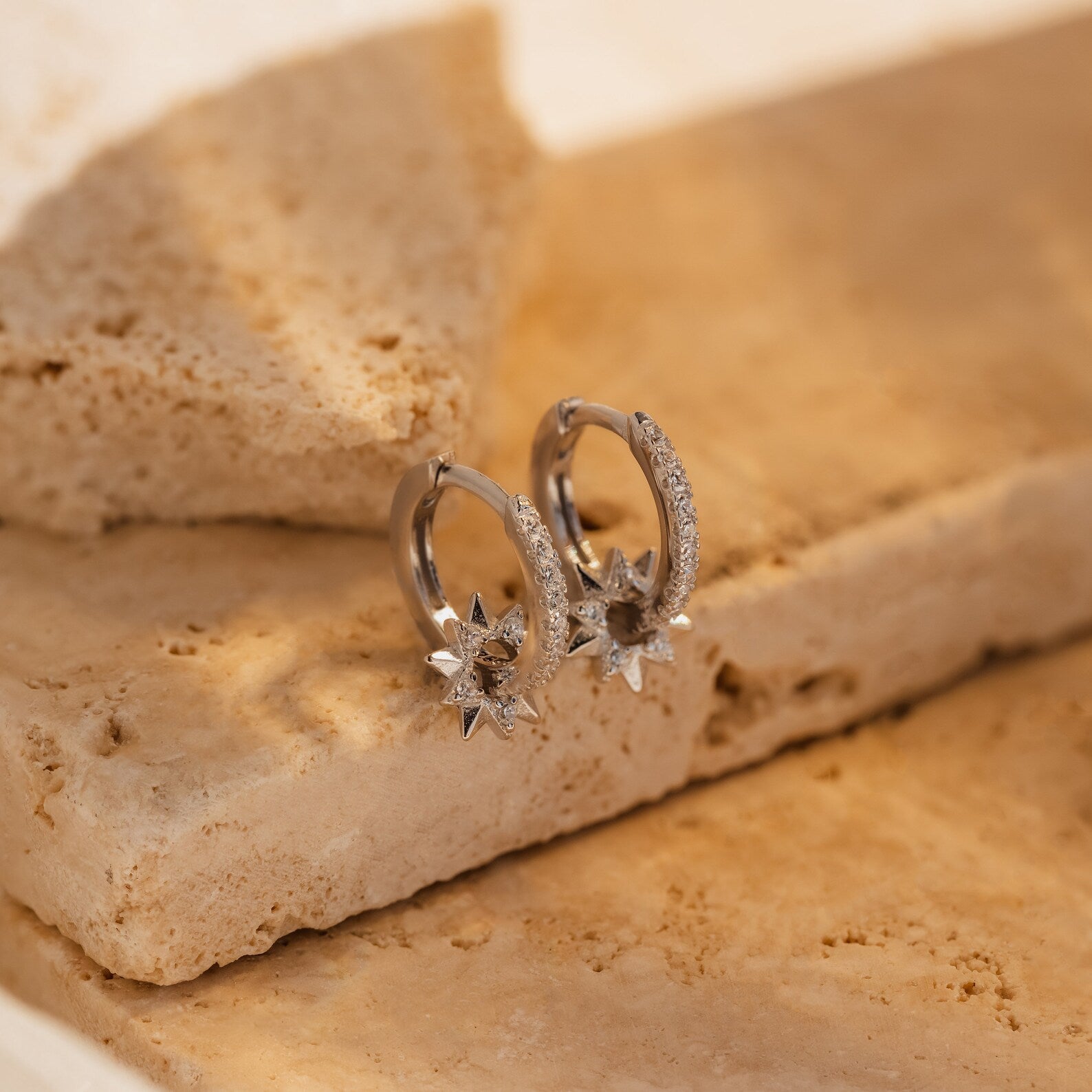 Pave Sun Huggies—silver hoop earrings with star-shaped pendants—are displayed on beige stone slabs in gentle, warm light.