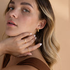 A woman with wavy blonde hair wears Bold Ribbed Hoops and rings, touching her face while dressed in a brown top.