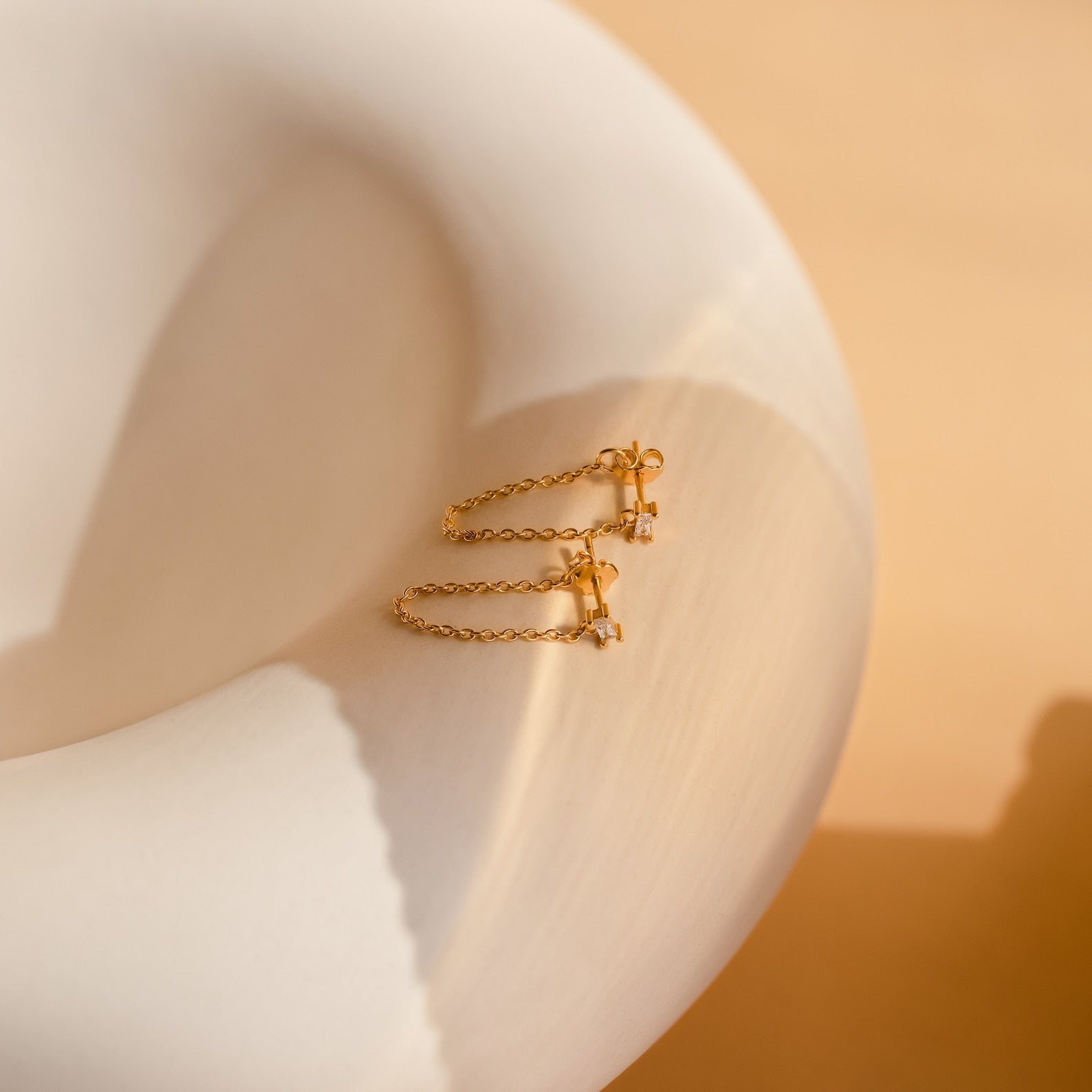 Baguette Chain Studs, featuring gold chains and small crystals ideal for a minimalist stack, are displayed on a smooth, curved white surface bathed in warm light.