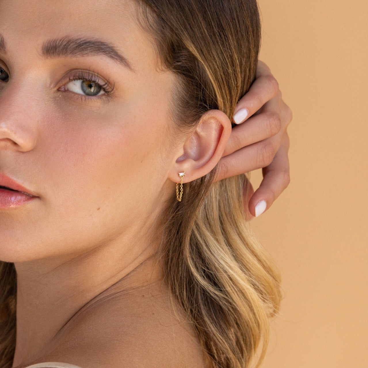 A woman with wavy blonde hair wears a small gold hoop from the Diamond Chain Earrings Set, gently touching her ear against a beige background.