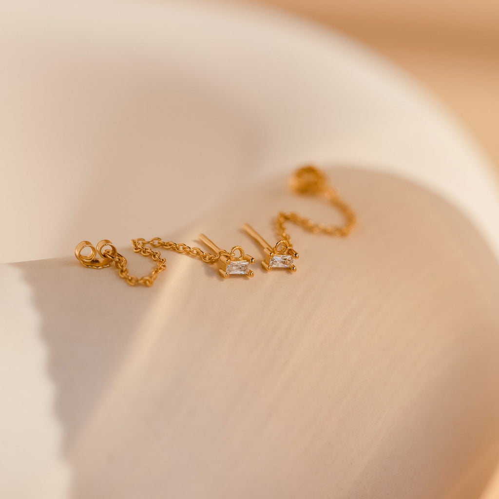 The Baguette Chain Studs—delicate gold earrings with small clear stones—rest on a curved beige surface, perfect for a minimalist stack.