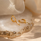 The Raine Opal Dagger Huggies, featuring opal and crystal details, are displayed on a textured stone surface with soft fabric nearby, channeling bohemian style.