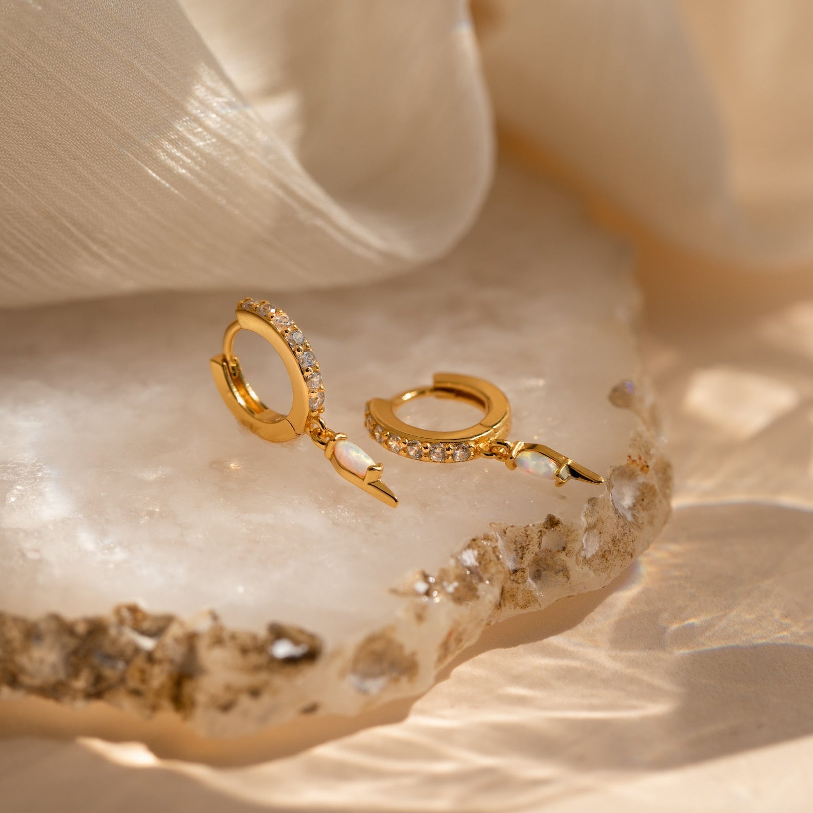 The Raine Opal Dagger Huggies, featuring opal and crystal details, are displayed on a textured stone surface with soft fabric nearby, channeling bohemian style.