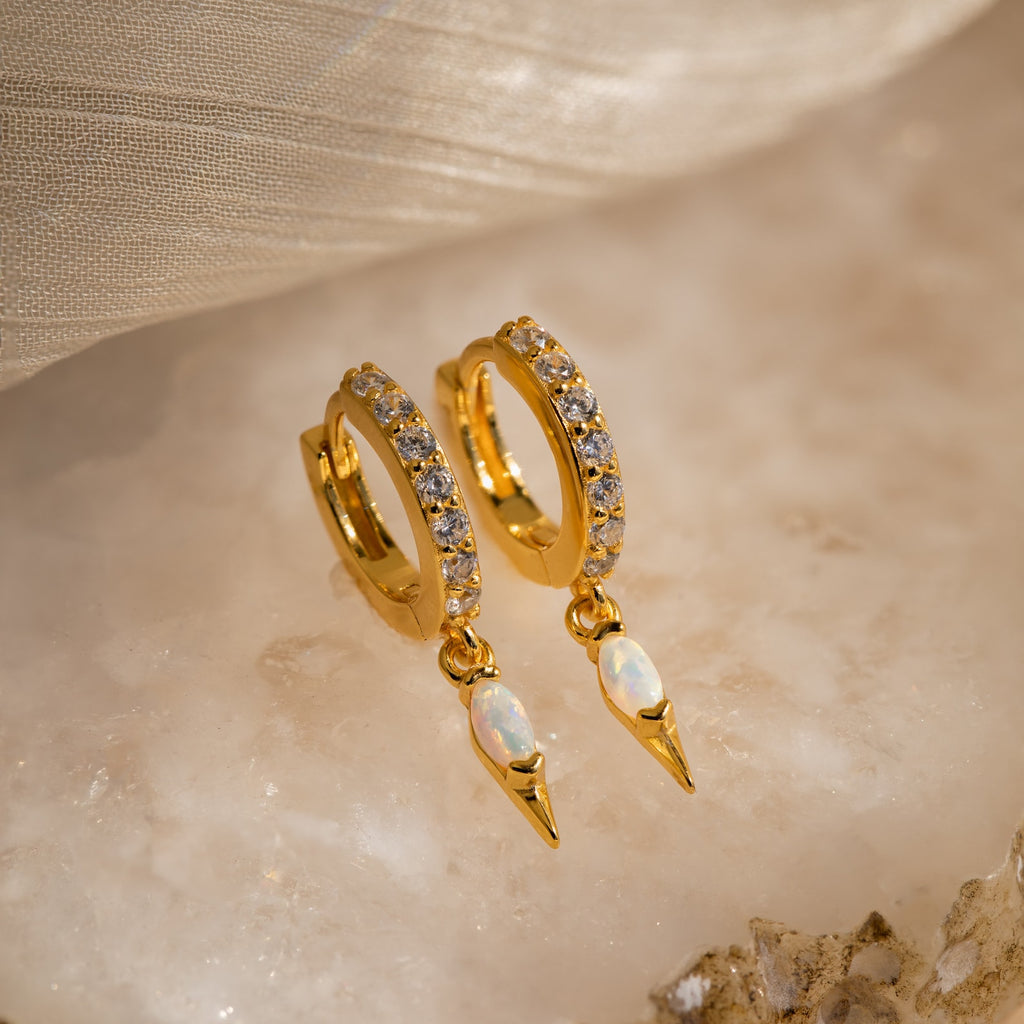 The Raine Opal Dagger Huggies feature gold hoops set with clear stones and opal teardrop pendants, adding a subtle sparkle and bohemian elegance to any outfit. Perfect for elevating your look with a free-spirited touch.