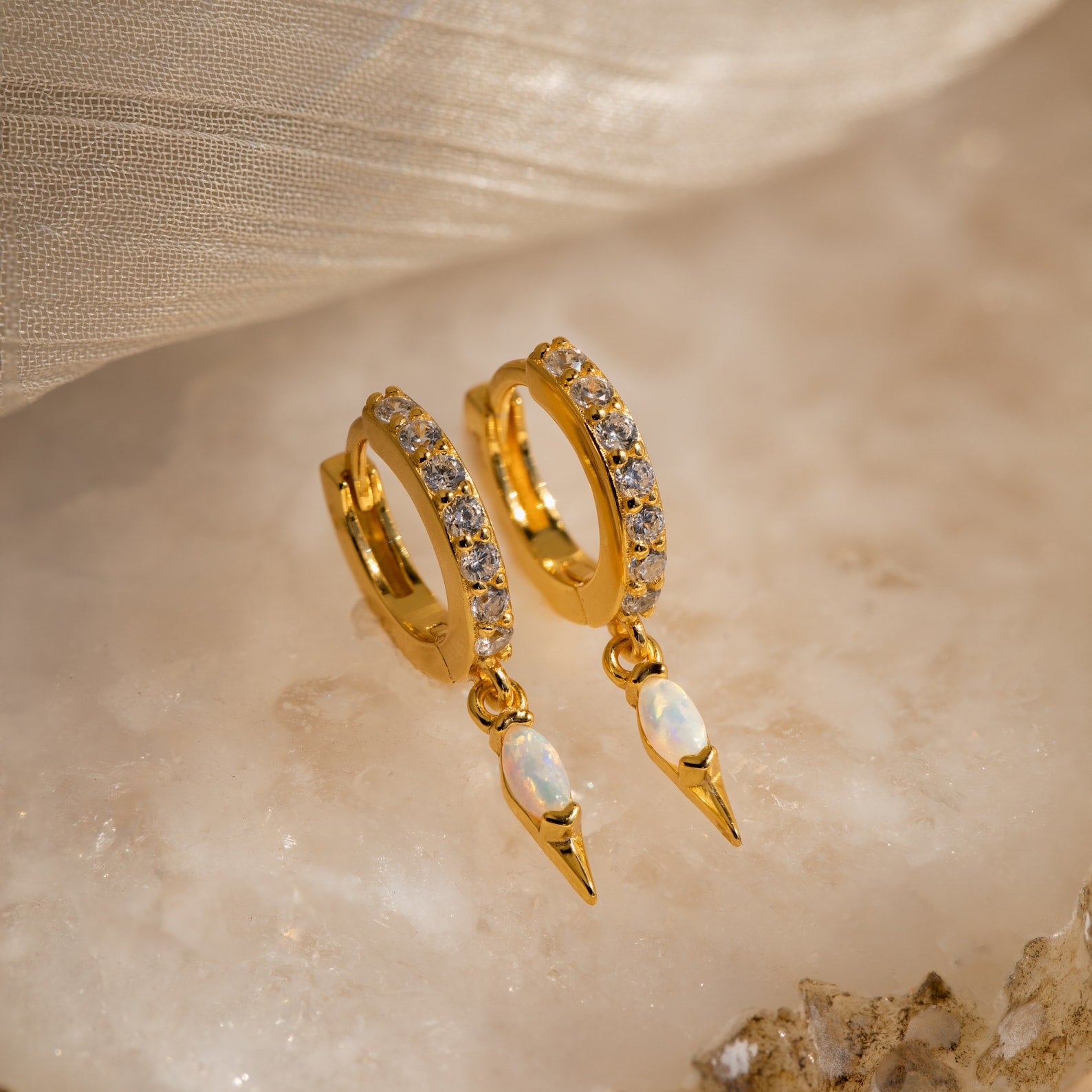 The Raine Opal Dagger Huggies feature gold hoops set with clear stones and opal teardrop pendants, adding a subtle sparkle and bohemian elegance to any outfit. Perfect for elevating your look with a free-spirited touch.