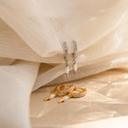 The Raine Opal Dagger Huggies, shown in silver and gold on soft beige fabric, add a subtle bohemian touch to any look with their unique design.