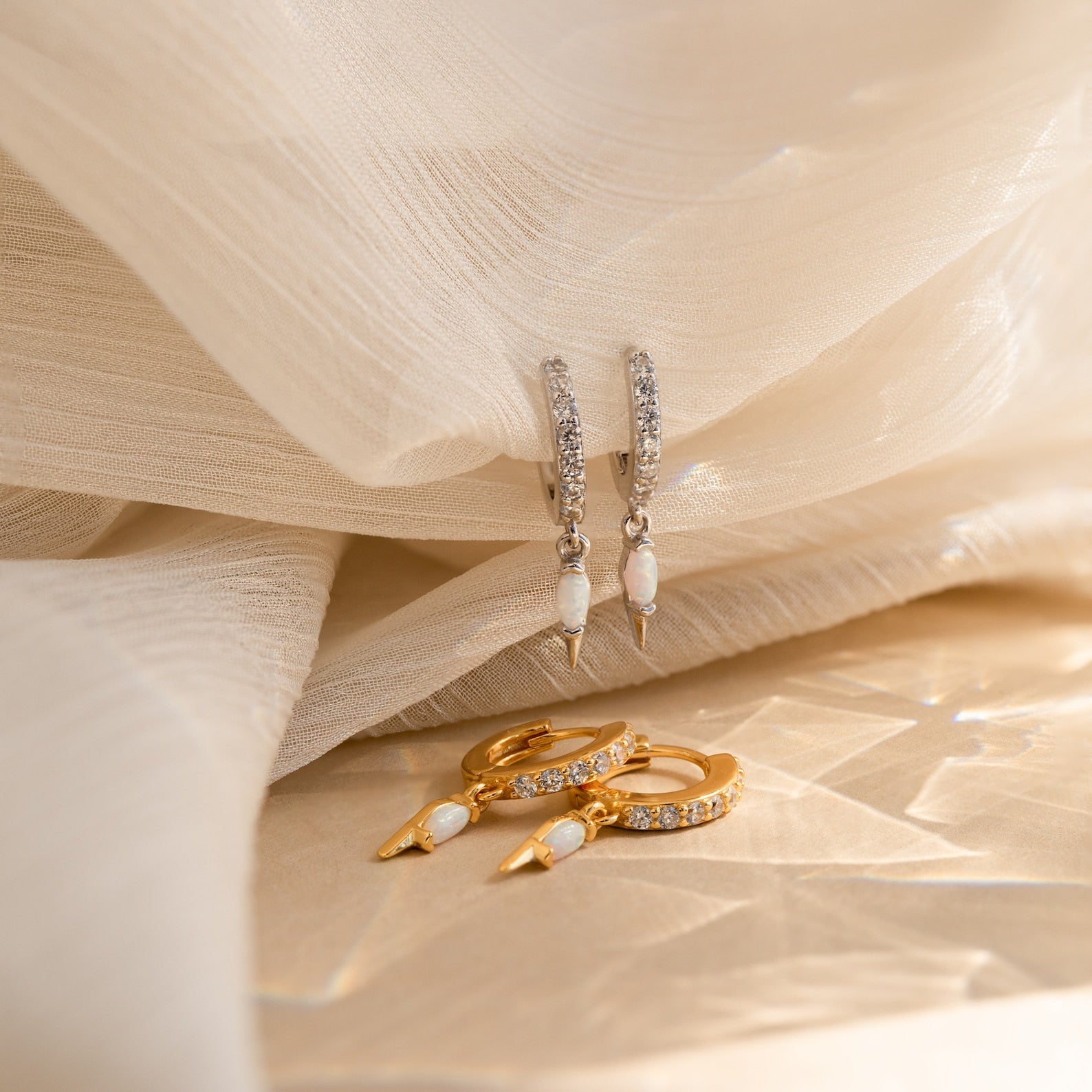 The Raine Opal Dagger Huggies, shown in silver and gold on soft beige fabric, add a subtle bohemian touch to any look with their unique design.