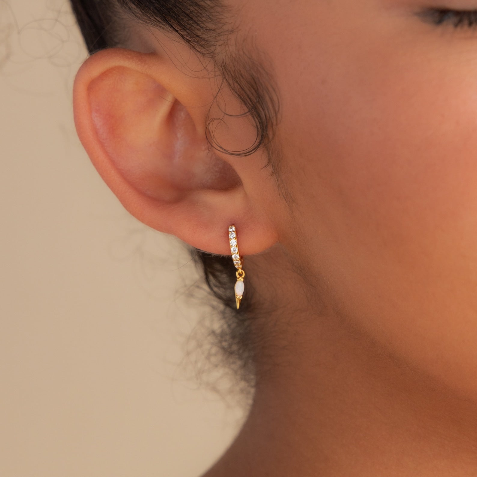 Close-up of a person's ear wearing the Raine Opal Dagger Huggies—small gold hoop earrings with opal gemstones and a dangling dagger charm, capturing a bohemian vibe.