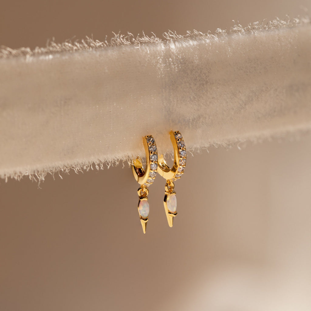 The Raine Opal Dagger Huggies, gold hoop earrings with dainty opals and dangling pearls, evoke a bohemian vibe and are elegantly displayed on a soft textured beige surface.