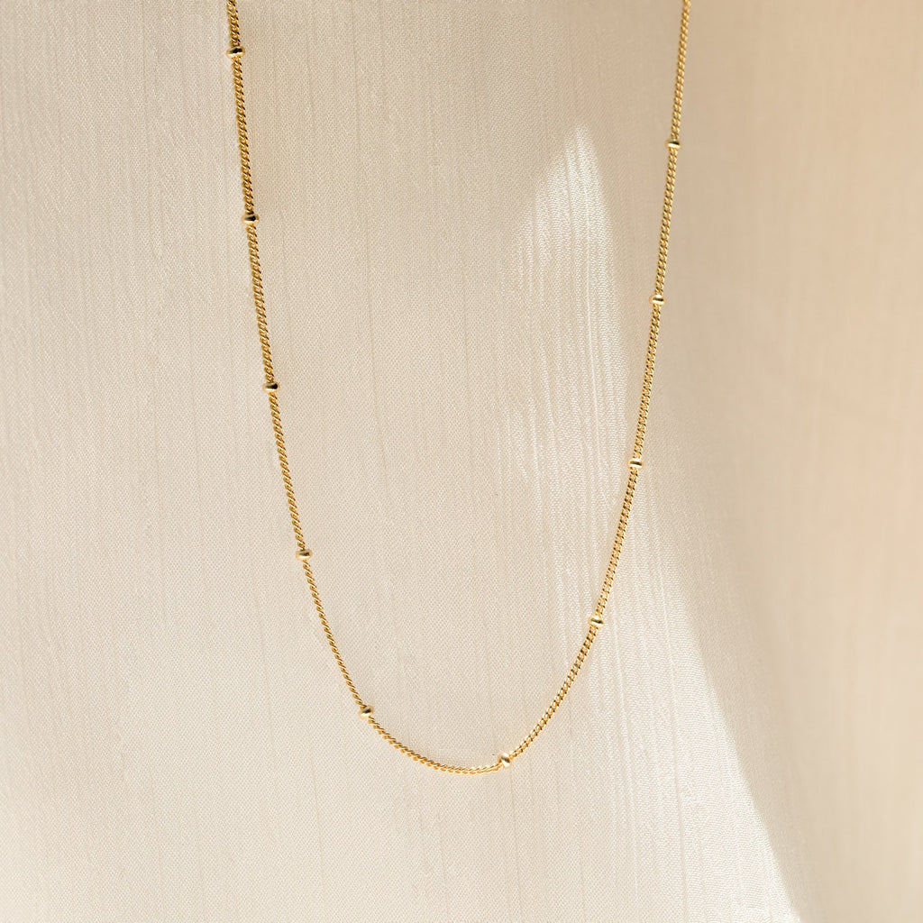 The Satellite Beaded Belly Chain features delicate gold with small bead accents, showcased against a soft beige fabric background.