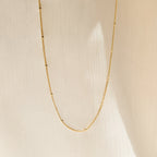 The Satellite Beaded Belly Chain features delicate gold with small bead accents, showcased against a soft beige fabric background.