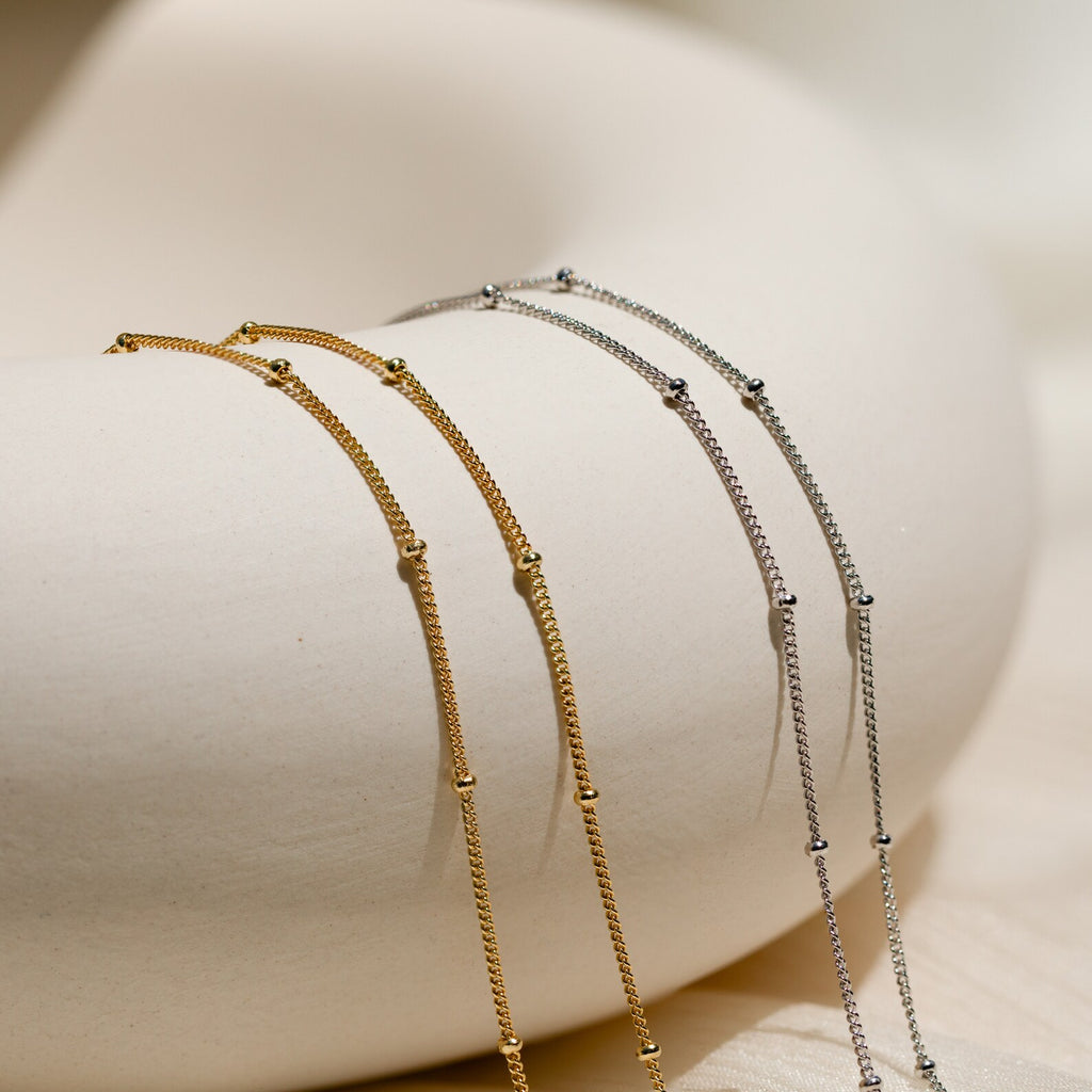 The Satellite Beaded Belly Chain features delicate gold and silver chains draped over a smooth, curved white surface, evoking understated sophistication with the elegance of a beaded waist chain.