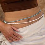 A close-up of a woman's waist adorned with the Satellite Beaded Belly Chain layered over a delicate belly chain and styled with a sheer white wrap.