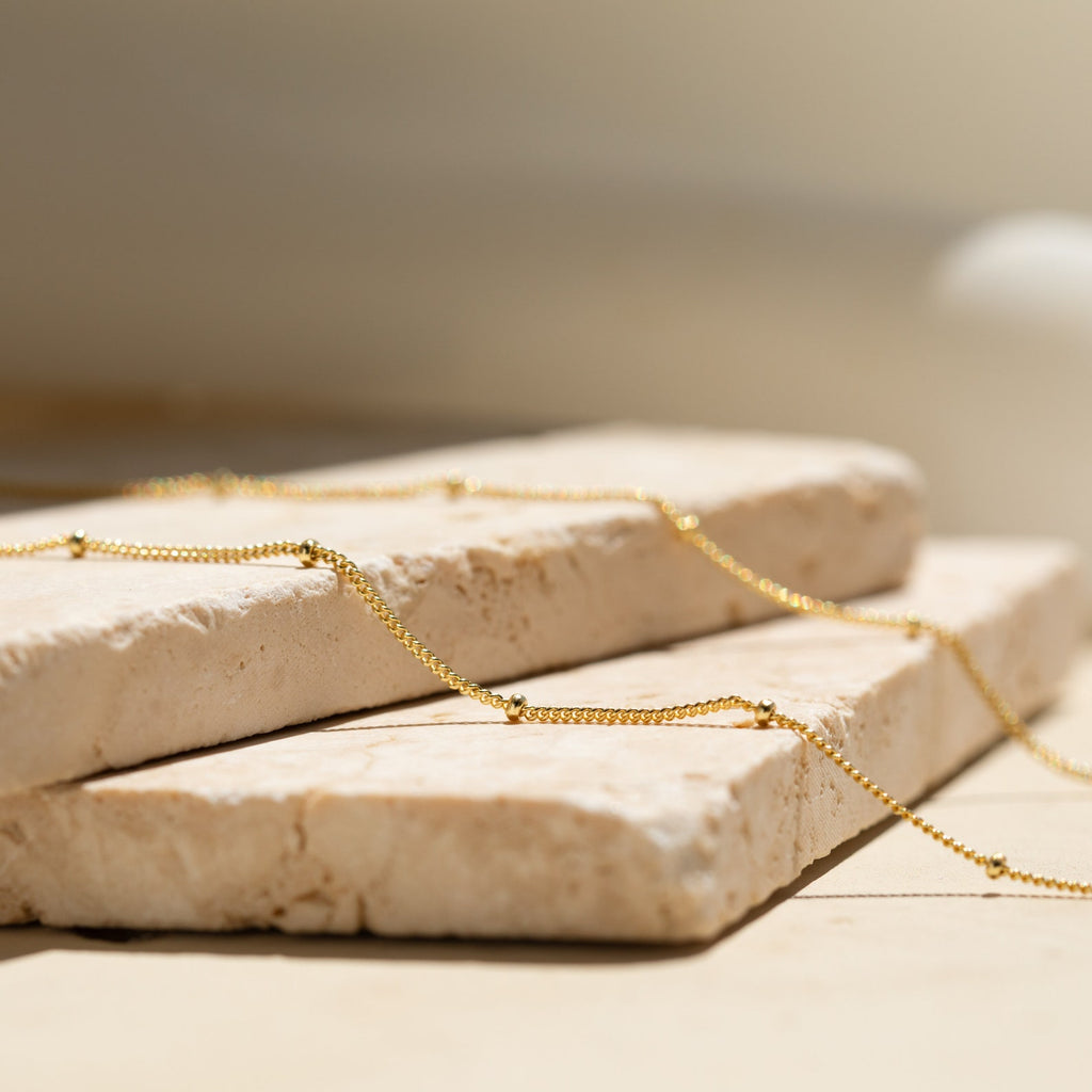 The Satellite Beaded Belly Chain, crafted in delicate gold, is showcased on two stacked beige stone slabs under soft natural light.