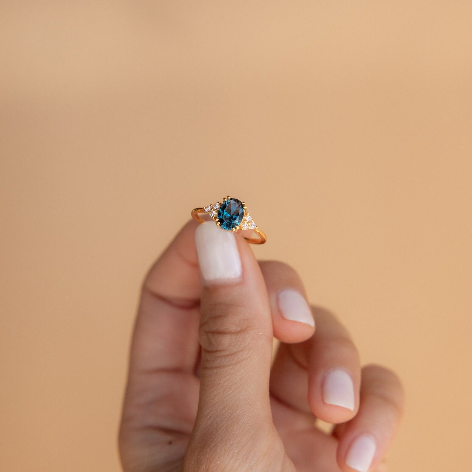 A hand with manicured nails holds the Elizabeth Blue Topaz Ring in 18K Gold, featuring a striking blue gemstone and glittering side stones in a vintage boho design.