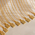 Gold initial pendant necklaces with rectangular charms featuring pavé crystal letters, displayed in a row on delicate chains over a soft fabric background.