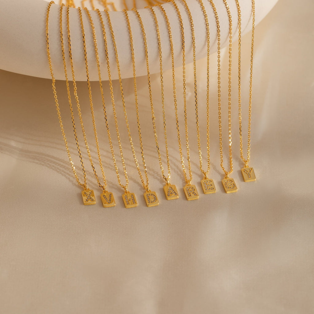 Mini Pave Initial Tag Necklaces, each featuring a unique letter, are displayed in a row on beige fabric—perfect personalized jewelry to add a special touch to any look.
