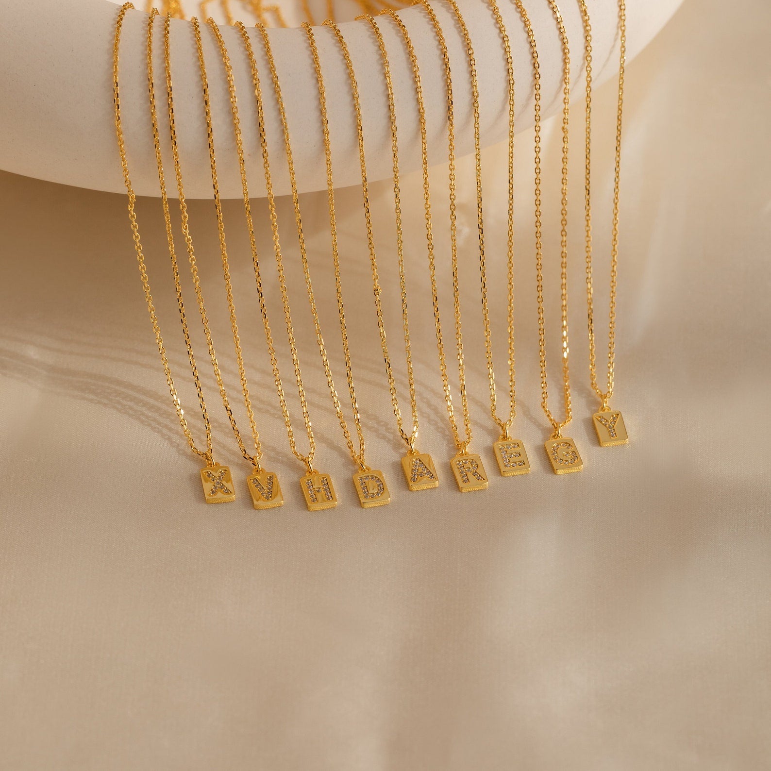 Mini Pave Initial Tag Necklaces, each featuring a unique letter, are displayed in a row on beige fabric—perfect personalized jewelry to add a special touch to any look.