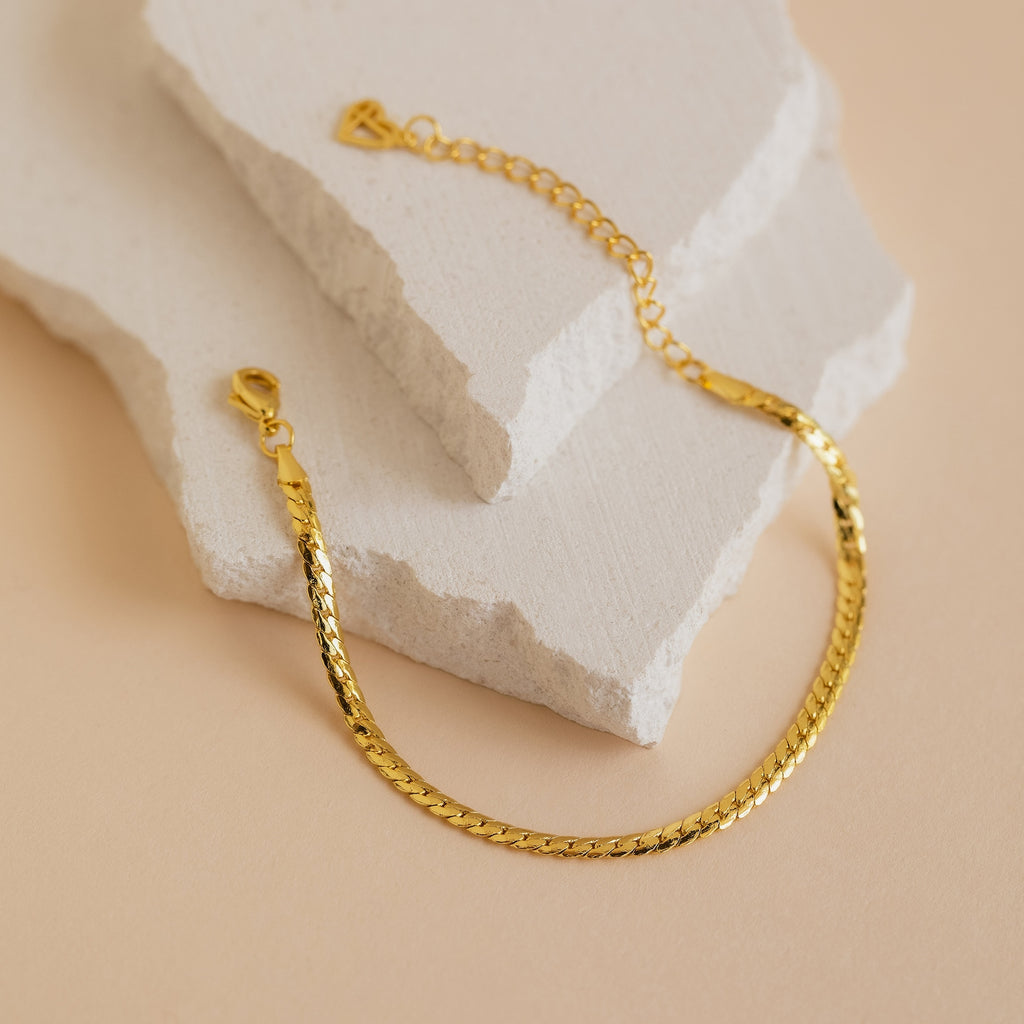 The Snake Chain Bracelet features a gold minimalist design with a lobster clasp, displayed on textured, off-white stone slabs against a beige background—ideal for layering into your bracelet stack.