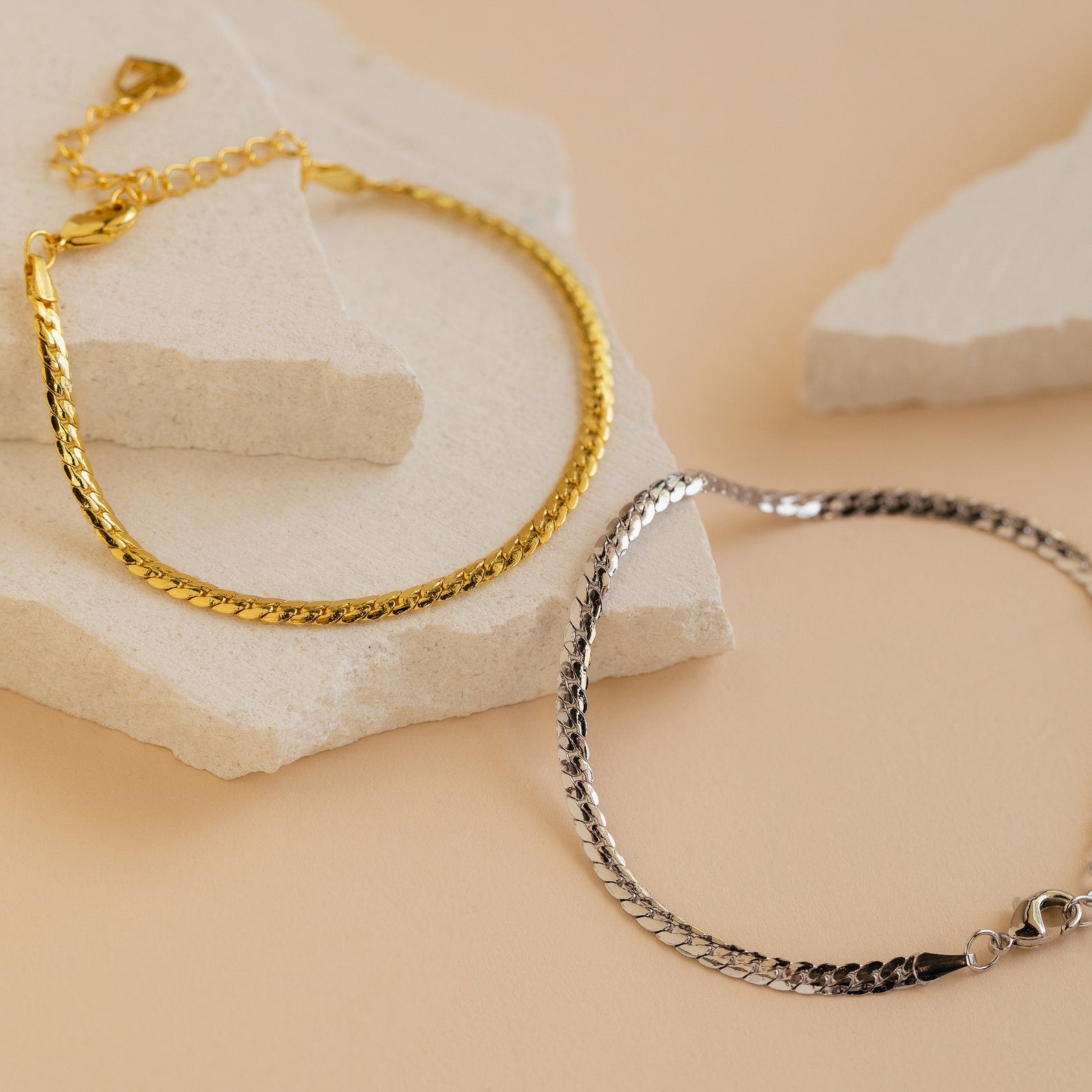 The Snake Chain Bracelet, a minimalist gold and silver accessory, is showcased on textured white stone slabs against a beige background.