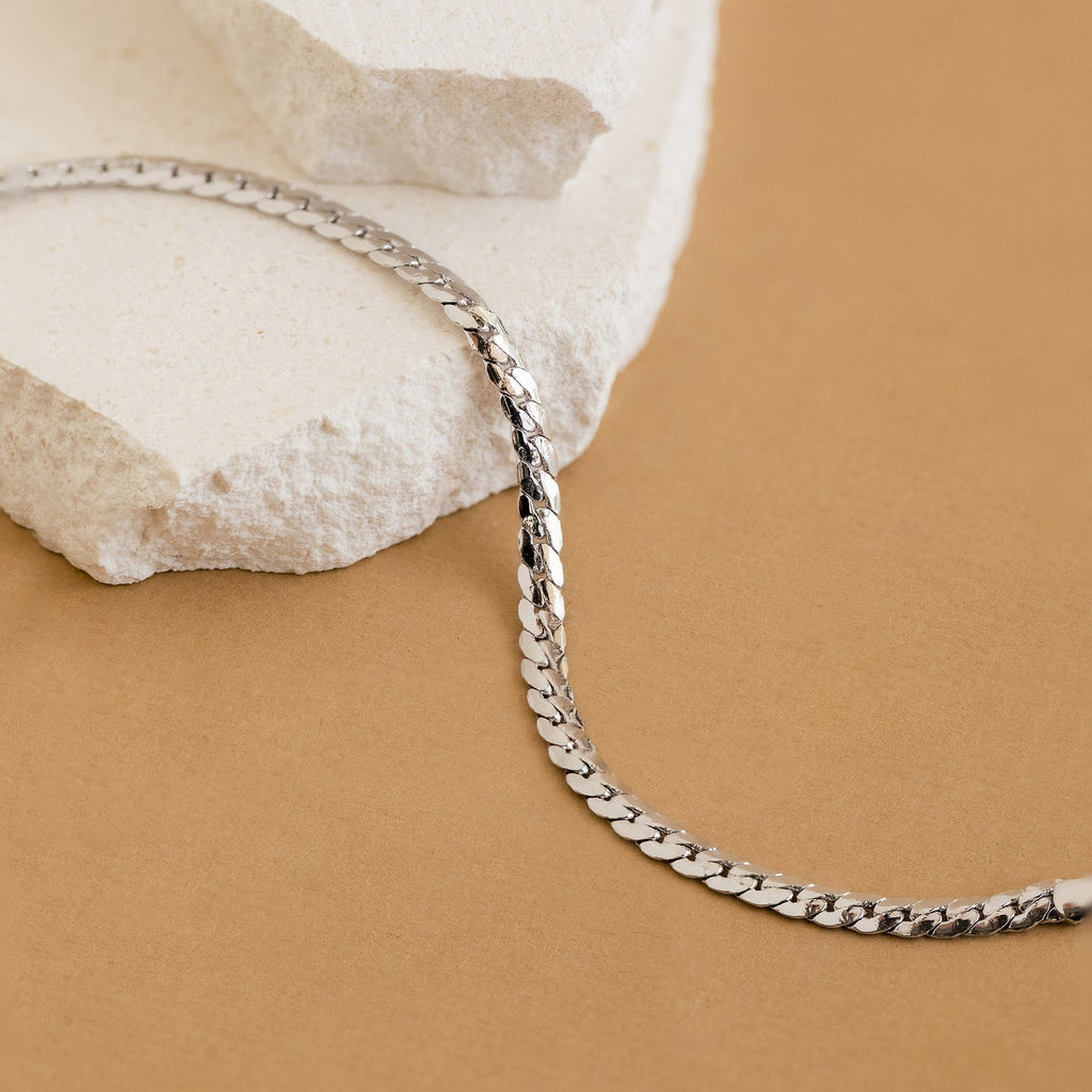 The Snake Chain Bracelet, ideal for minimalist stacks, is shown on white stone with a tan background.