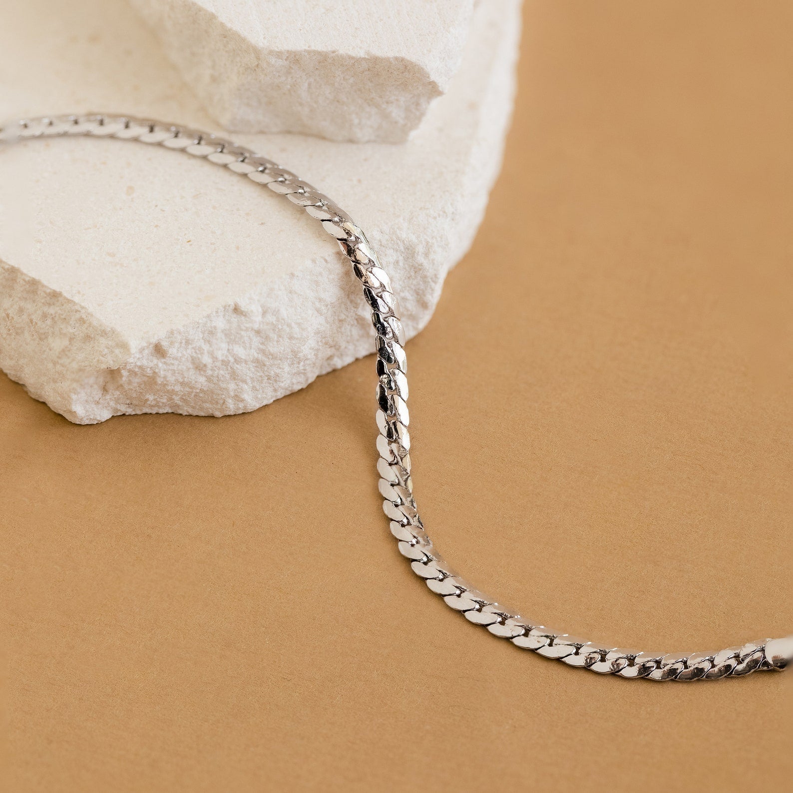 The Snake Chain Bracelet, ideal for minimalist stacks, is shown on white stone with a tan background.