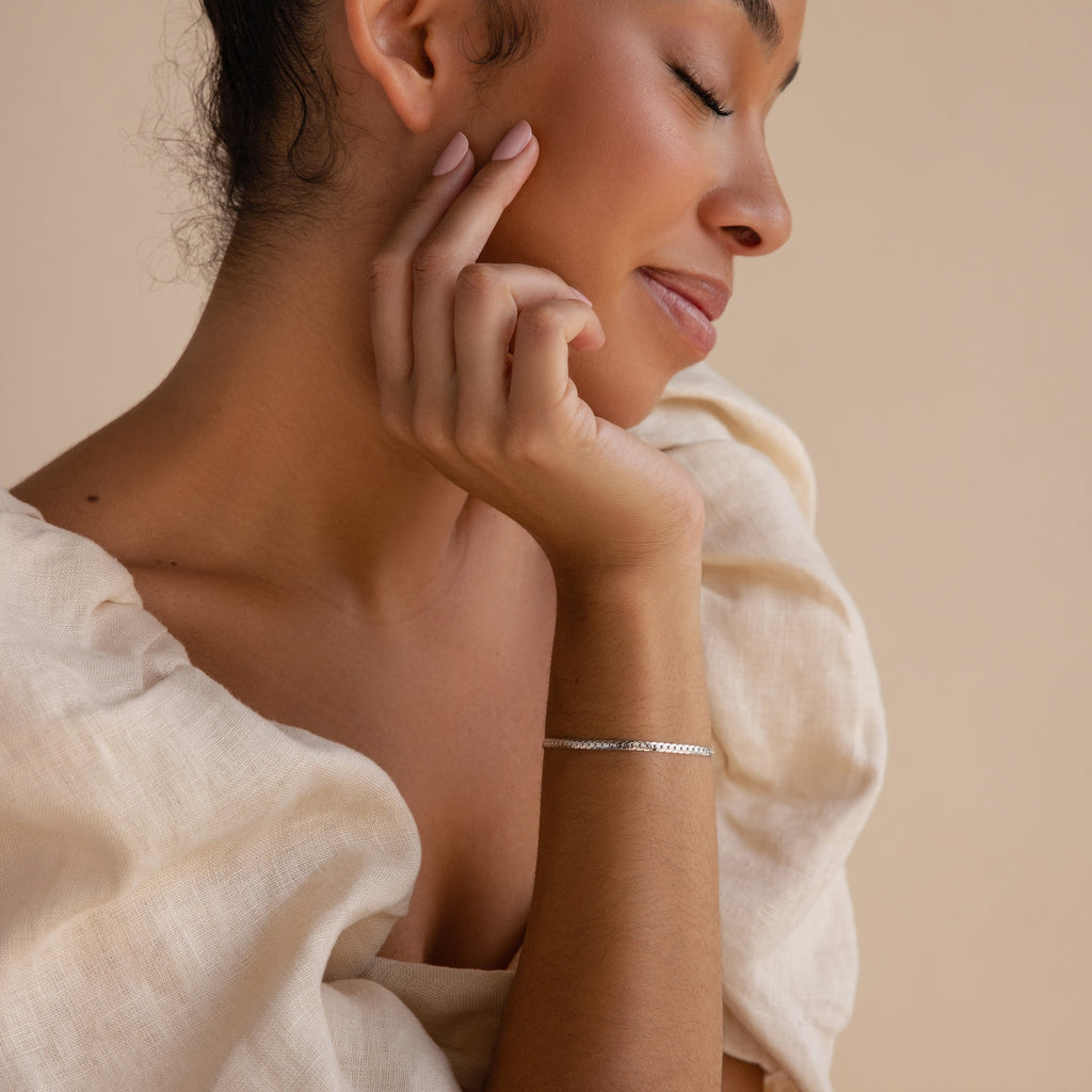 A woman with closed eyes gently touches her face, dressed in a cream blouse and wearing a delicate bracelet stack that highlights the sleek Snake Chain Bracelet.