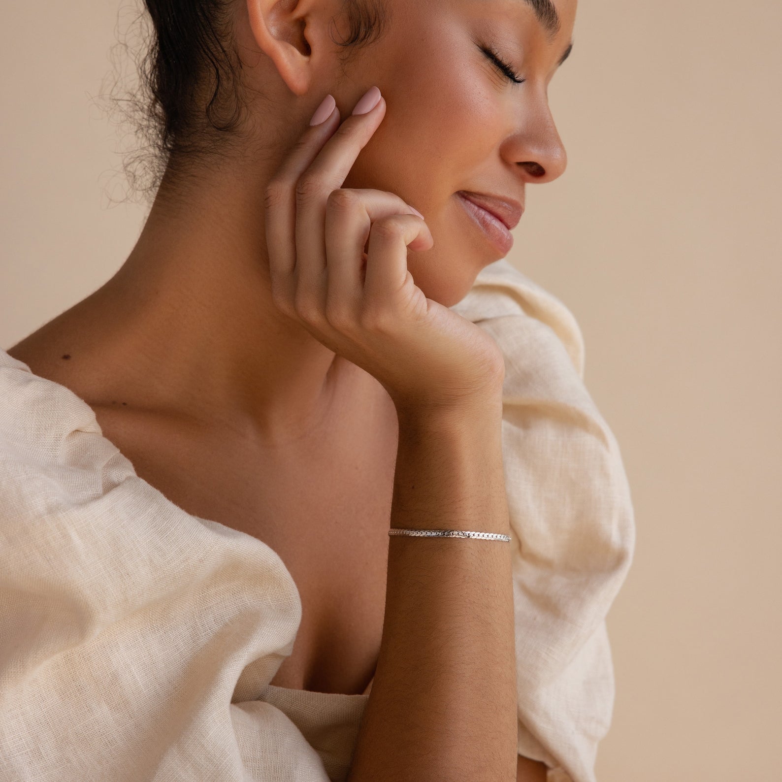 A woman with closed eyes gently touches her face, dressed in a cream blouse and wearing a delicate bracelet stack that highlights the sleek Snake Chain Bracelet.