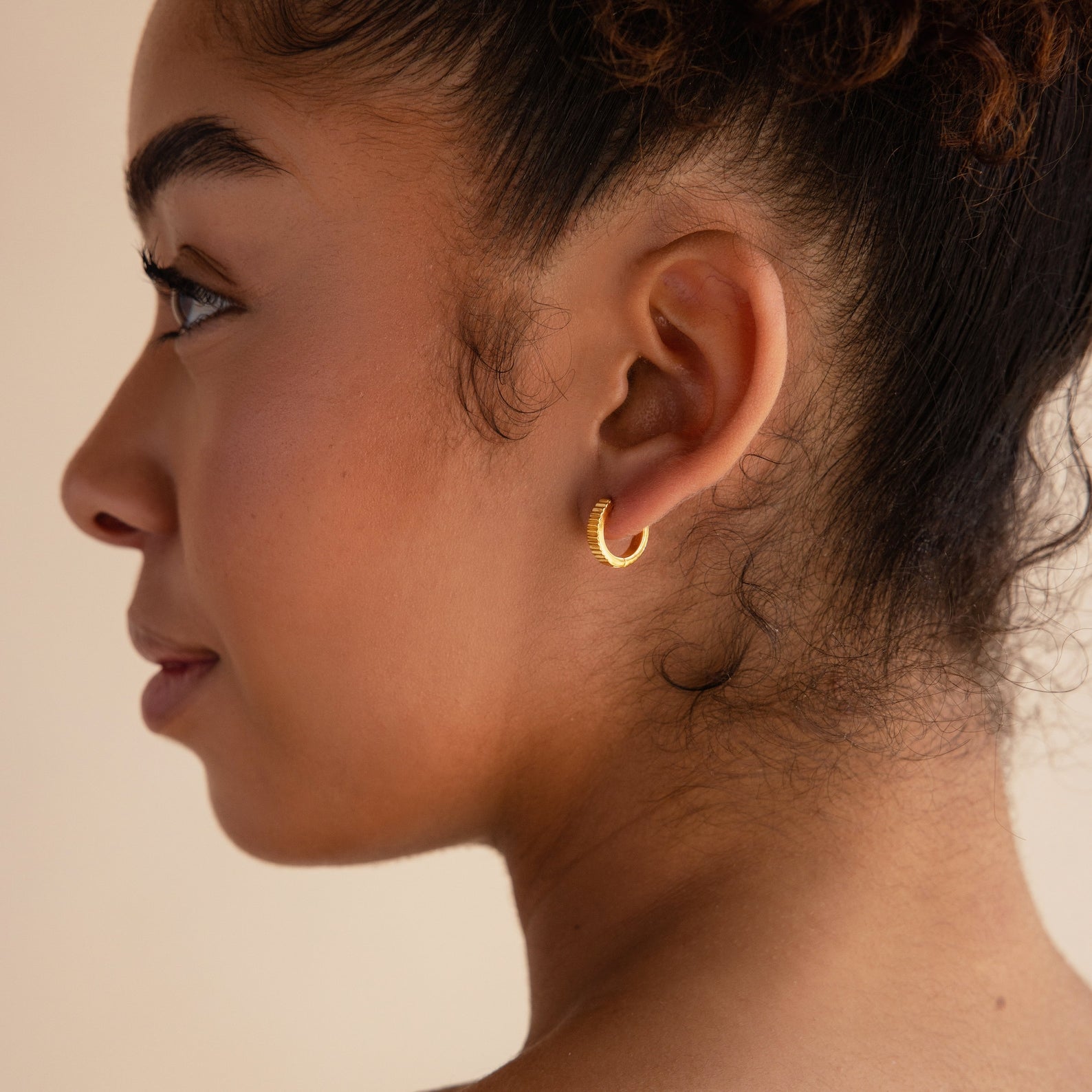 A woman in profile wears Ribbed Huggies, her curly hair pulled back to showcase the dainty earrings against a neutral background.