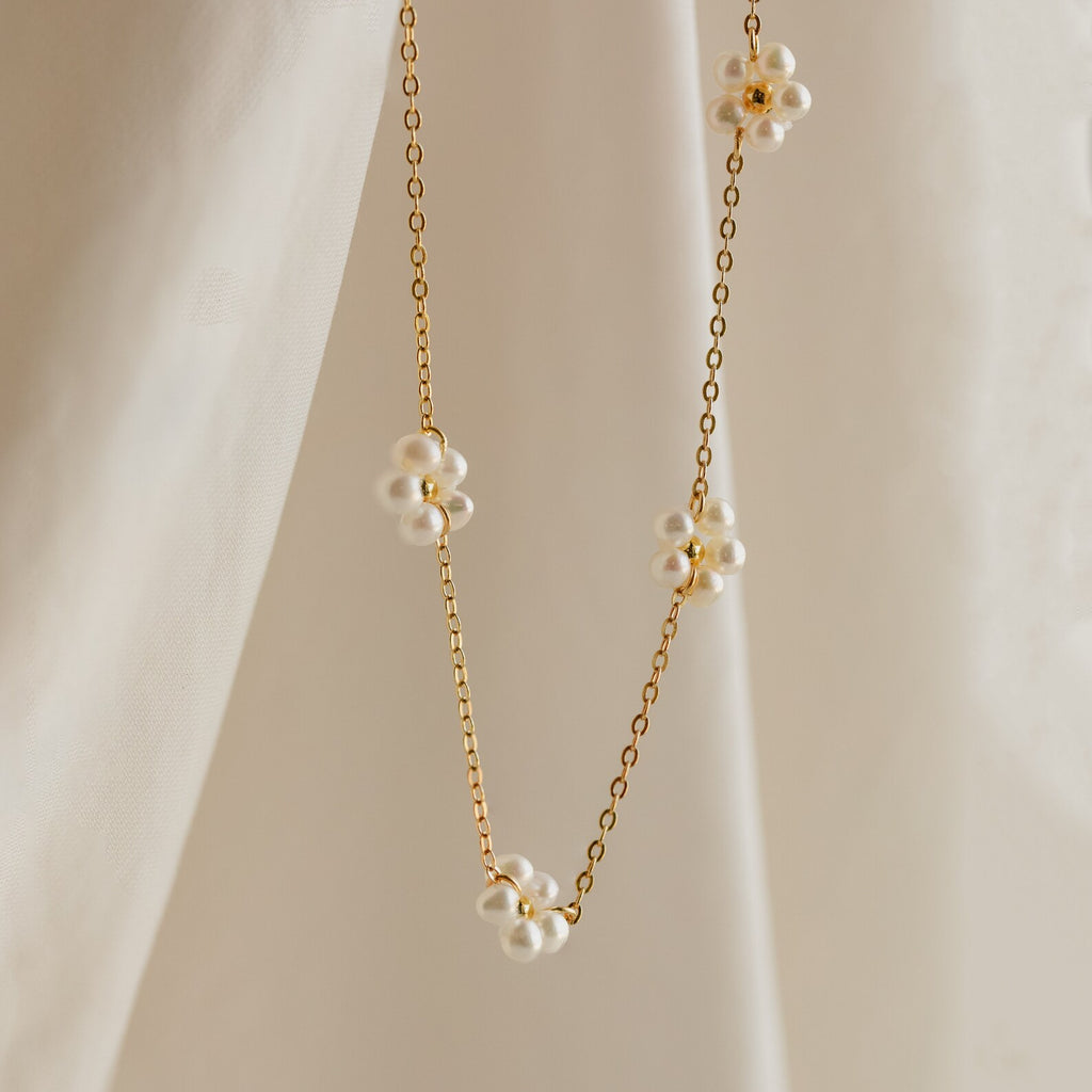 The Flower Pearl Station Necklace in 18K Gold features delicate flower-shaped pearl clusters, shown draped on soft beige fabric—a perfect bridesmaids gift or timeless accessory.
