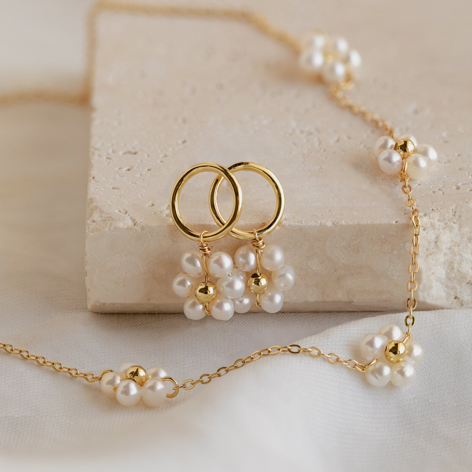 The Flower Pearl Station Necklace, displayed with gold hoop earrings on a beige stone slab, makes an elegant bridesmaids or Mother’s Day gift.