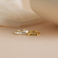 A Thin Name Signet Ring in silver with "Nova" and a gold ring featuring "Oliver" and a small gem are displayed on a soft beige surface.