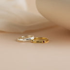 A Thin Name Signet Ring in silver with "Nova" and a gold ring featuring "Oliver" and a small gem are displayed on a soft beige surface.