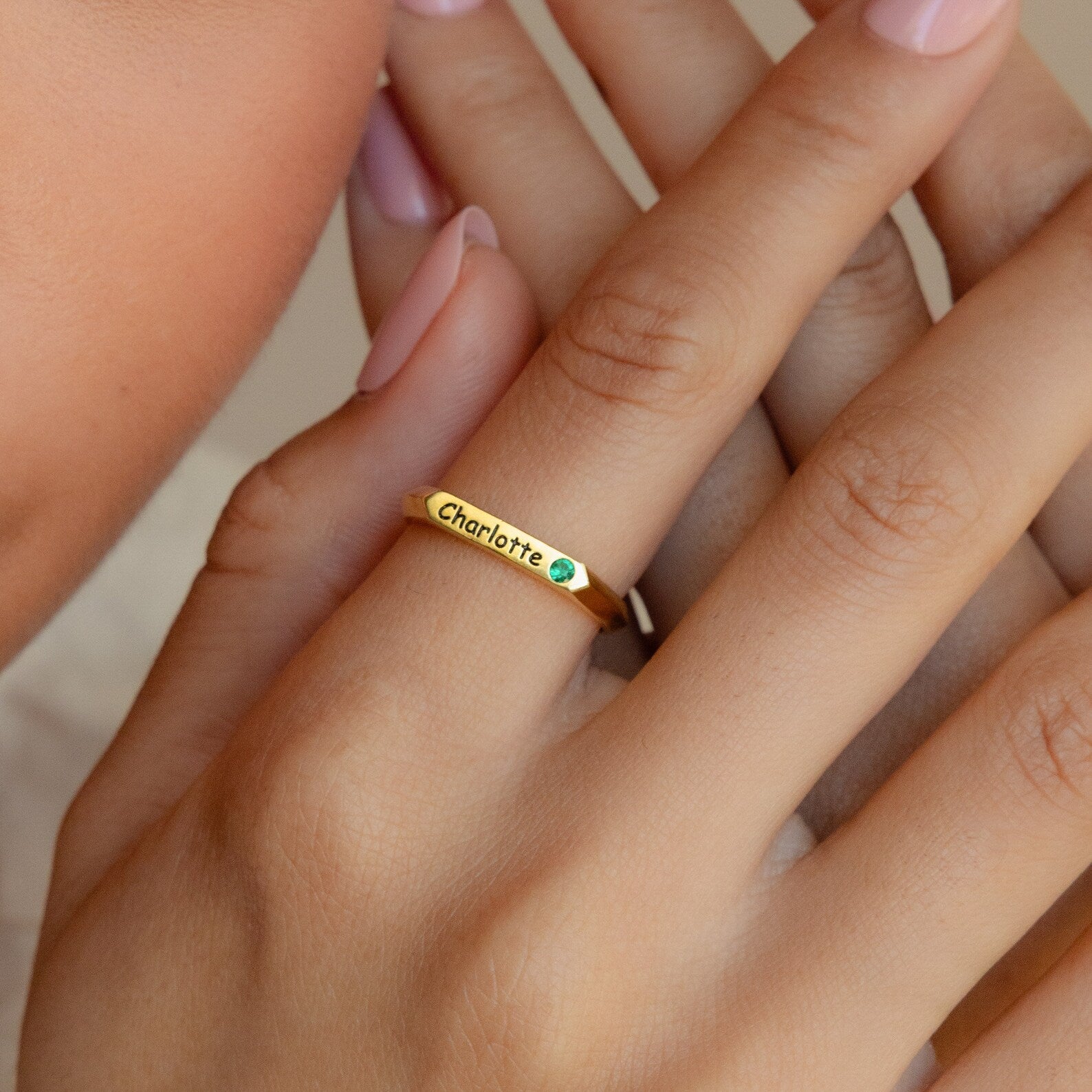 The Thin Name Signet Ring engraved with "Charlotte" features a small green gem, shining on a manicured hand—perfect for stacking or wearing solo in classic signet style.