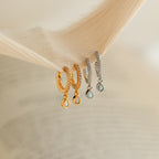 Two pairs of drop earrings, including elegant Opal Drop Huggies in gold and silver, displayed on the edge of a textured fabric.