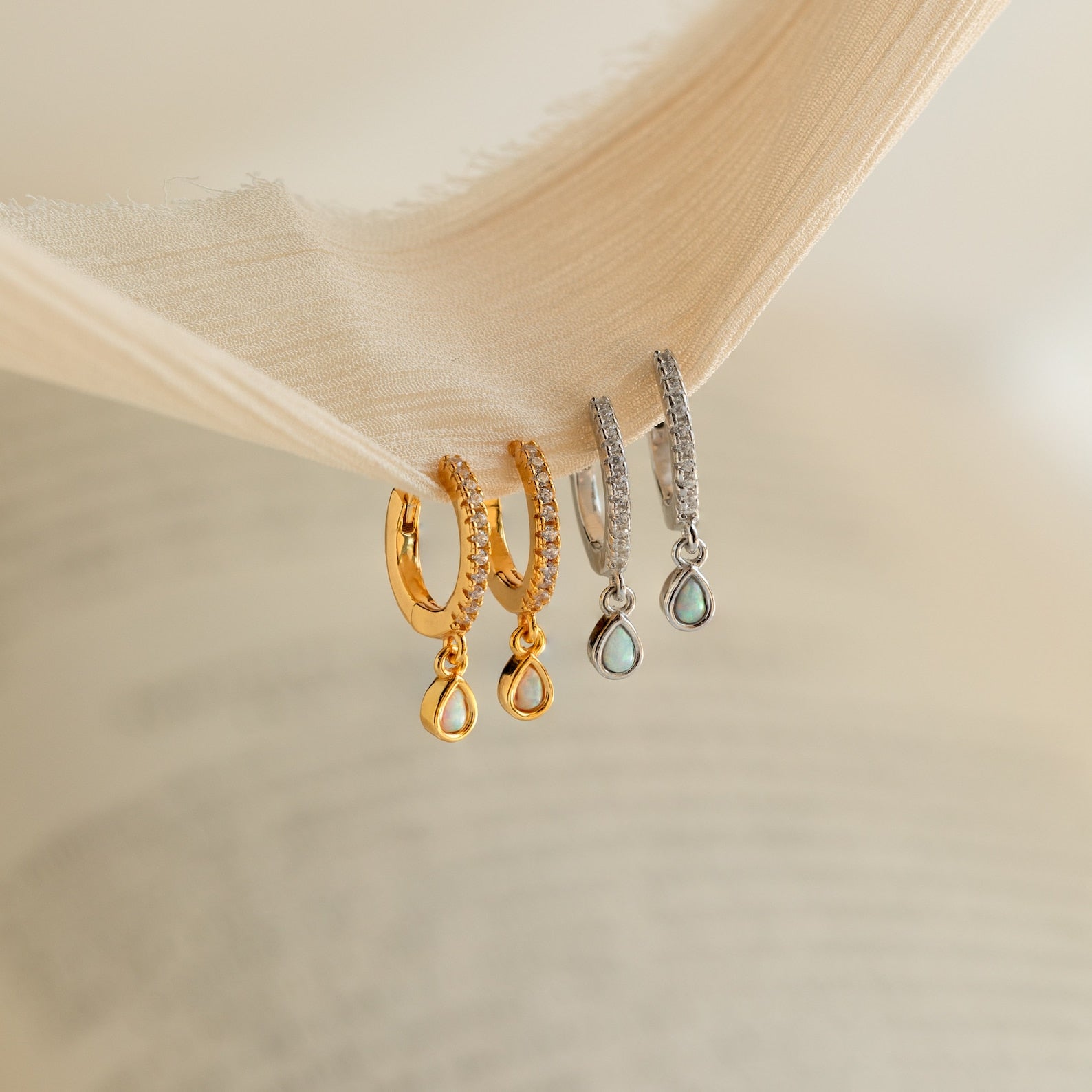 Two pairs of drop earrings, including elegant Opal Drop Huggies in gold and silver, displayed on the edge of a textured fabric.