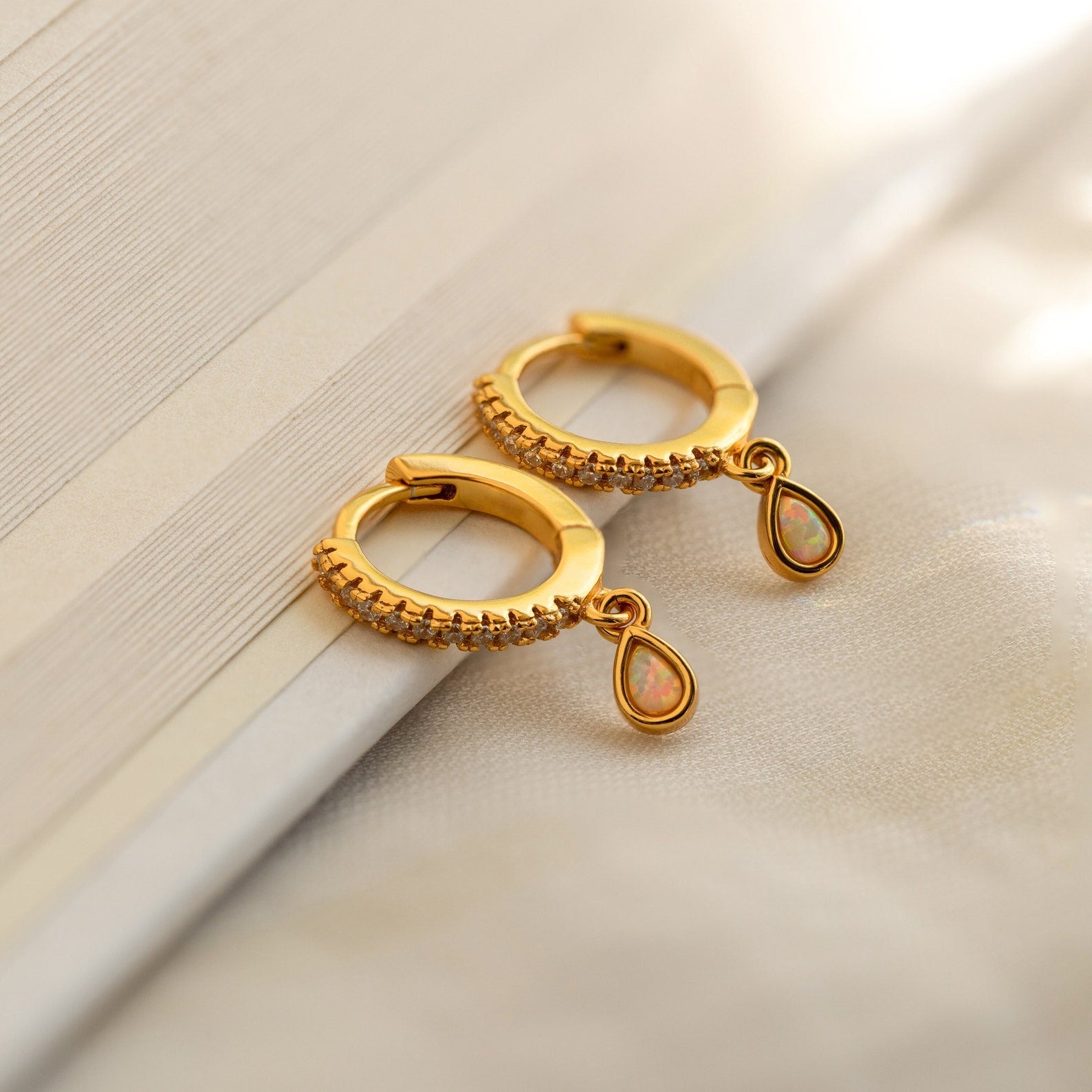 Opal Drop Huggies feature gold hoops, small white stones, and teardrop-shaped opal charms resting elegantly on an open book.