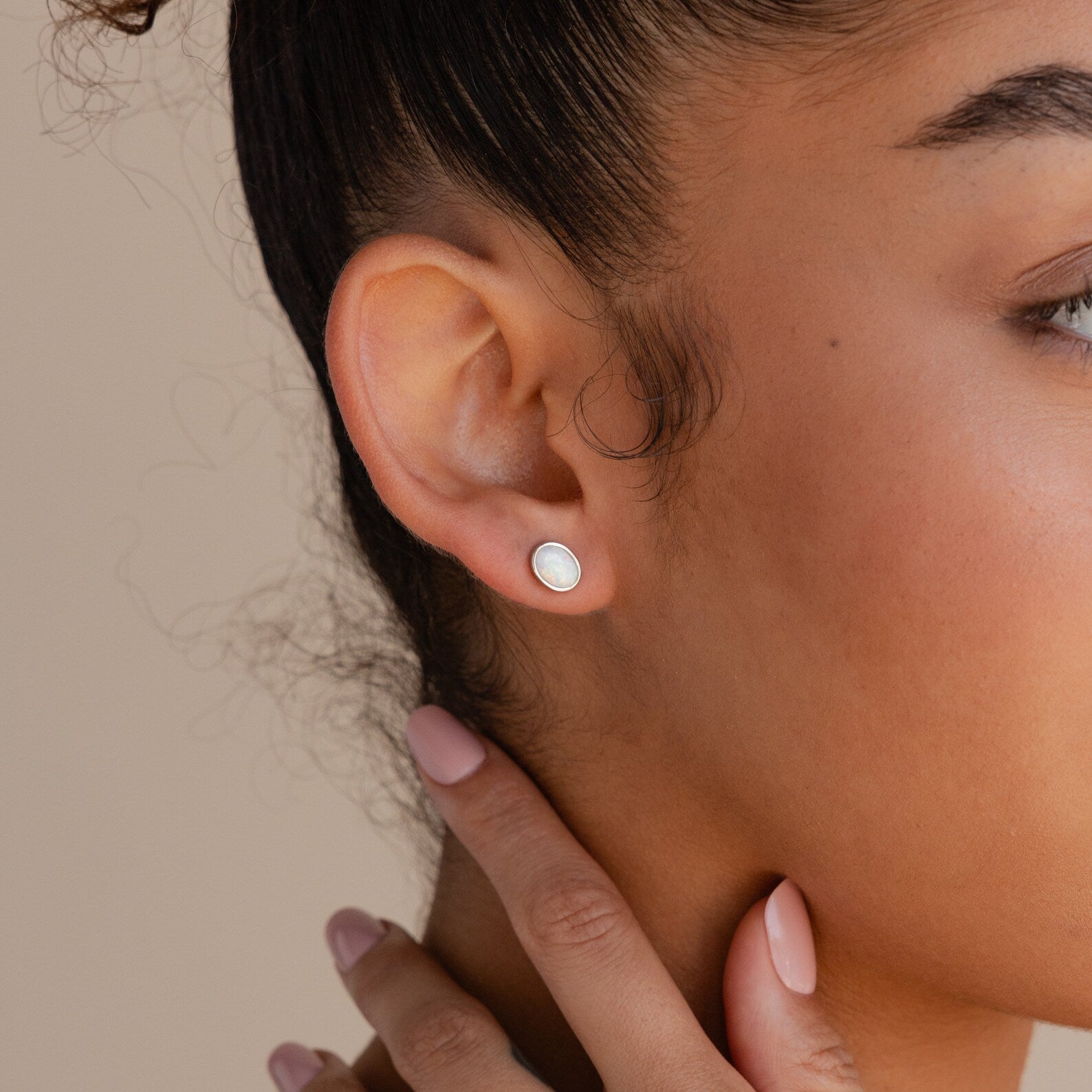 Close-up of a woman's ear wearing Oval Opal Studs, elegant earrings featuring a luminous white opal. Her hand touches her neck, nails painted light pink.