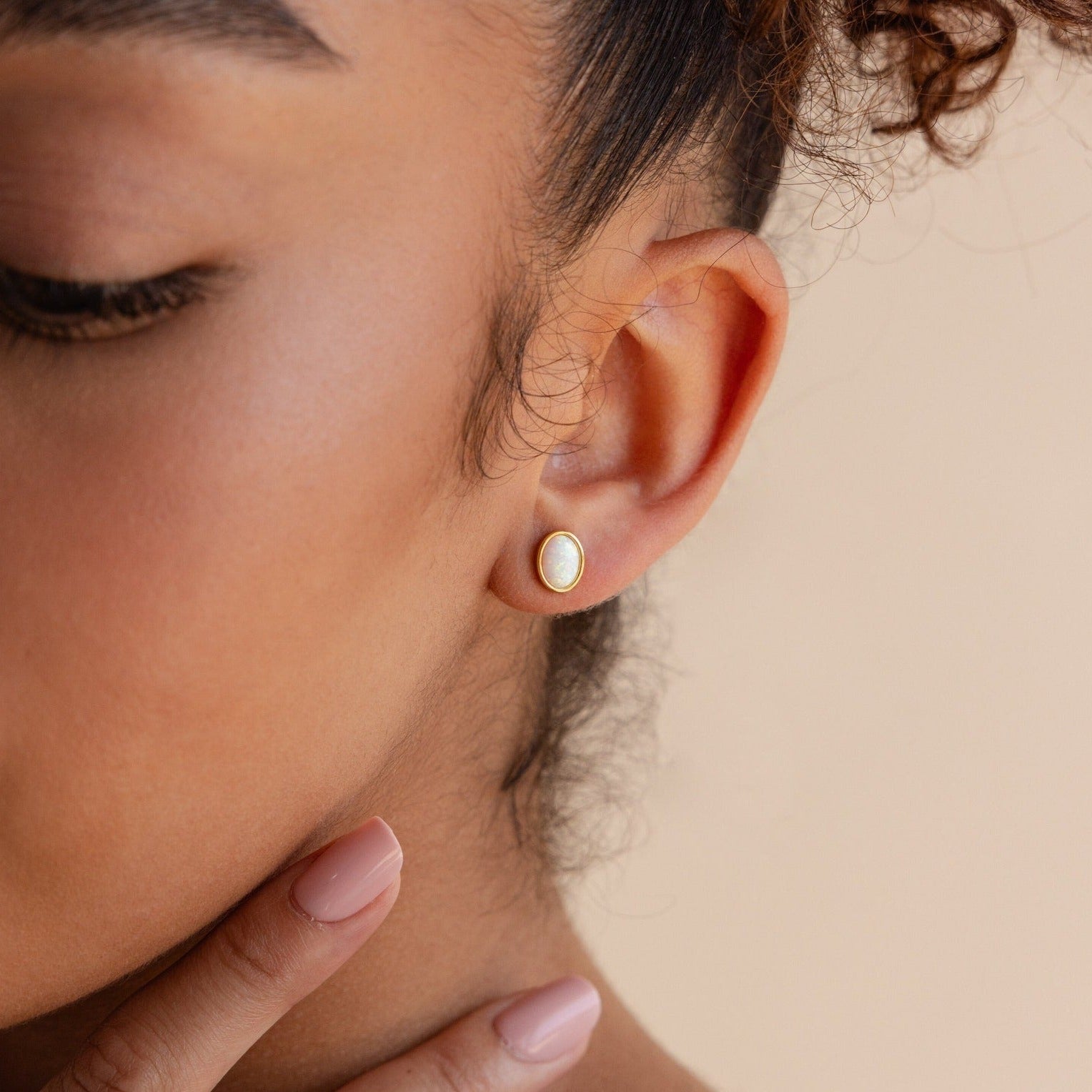 Close-up of a woman's ear wearing Oval Opal Studs, featuring an elegant white opal stone, as she gently touches her jawline.