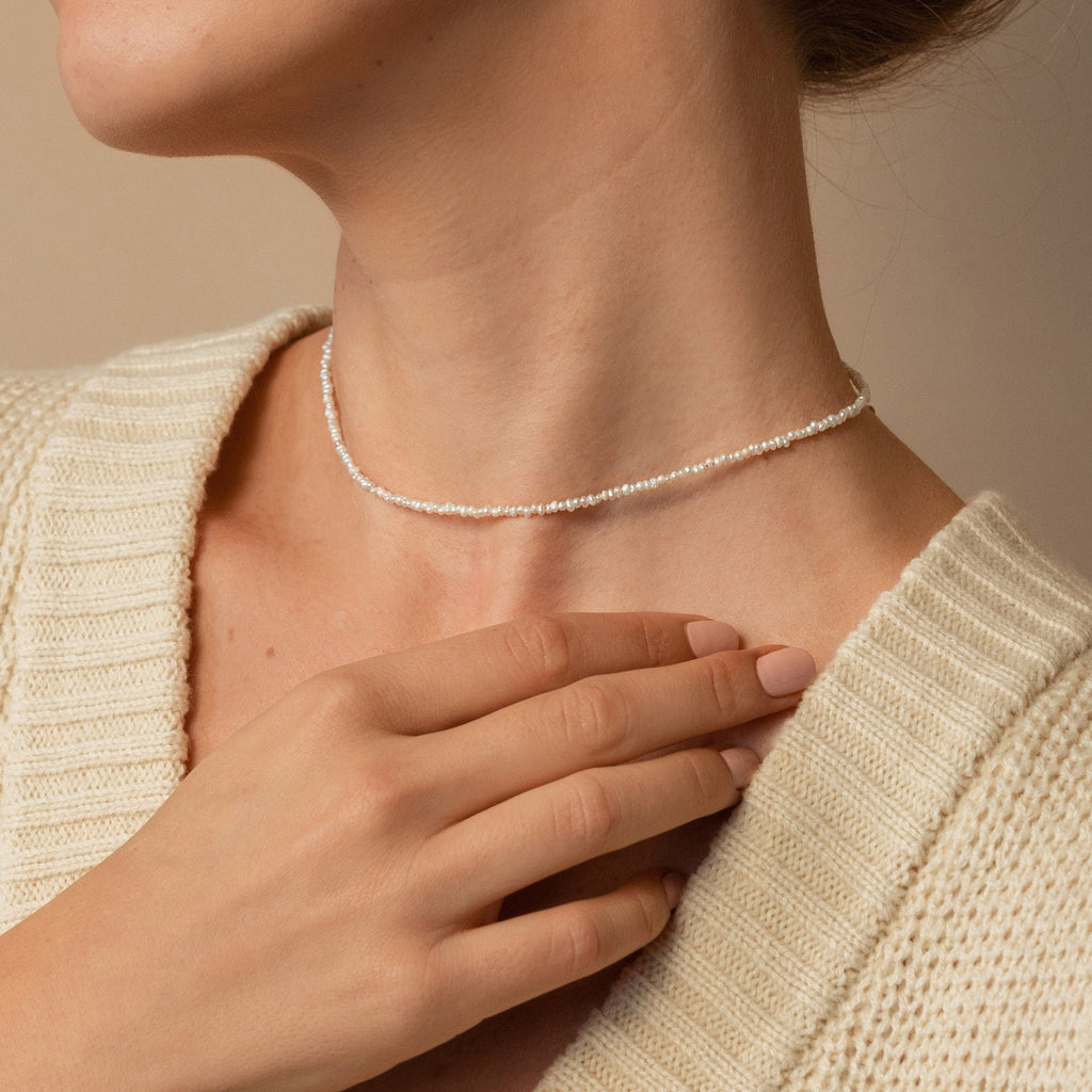 A woman in a cream knit sweater gently touches her neck, showcasing Boho Beaded Necklaces—a simple silver choker perfect for layering with summer outfits.