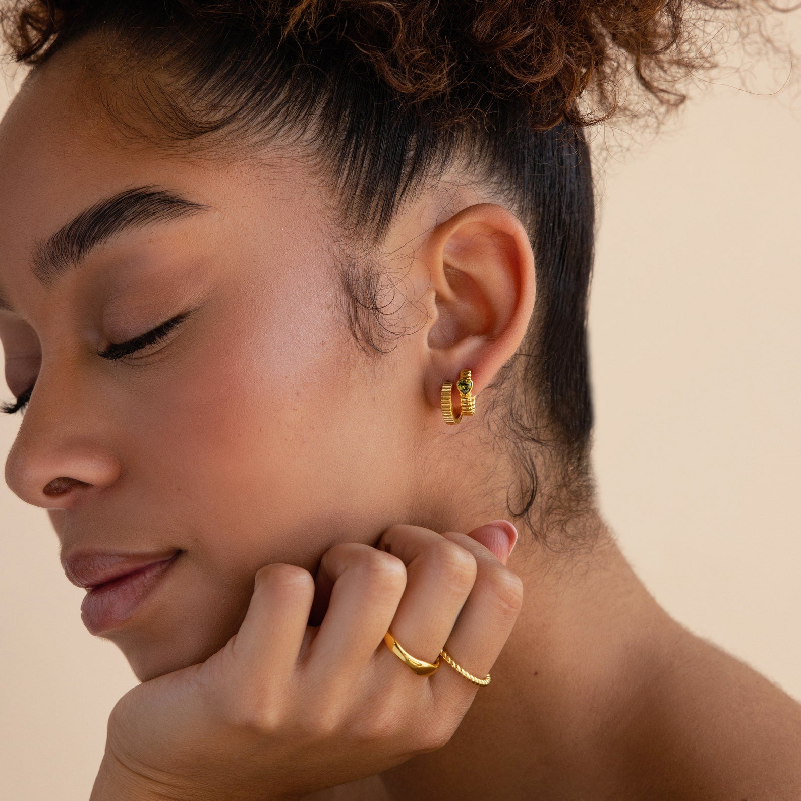 A woman with closed eyes wears Ribbed Huggies earrings and rings, resting her chin on her hand.