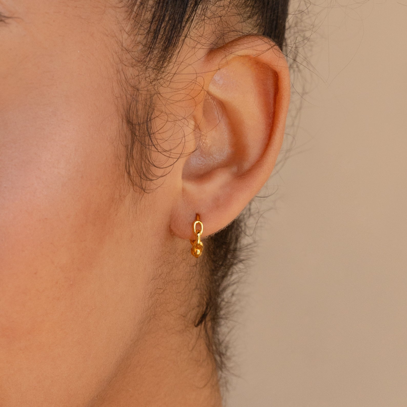 Close-up of a person's ear wearing Link Huggies, small gold hoop earrings with a dangling gold bead, paired with dainty chain earrings for an elegant earring stack.