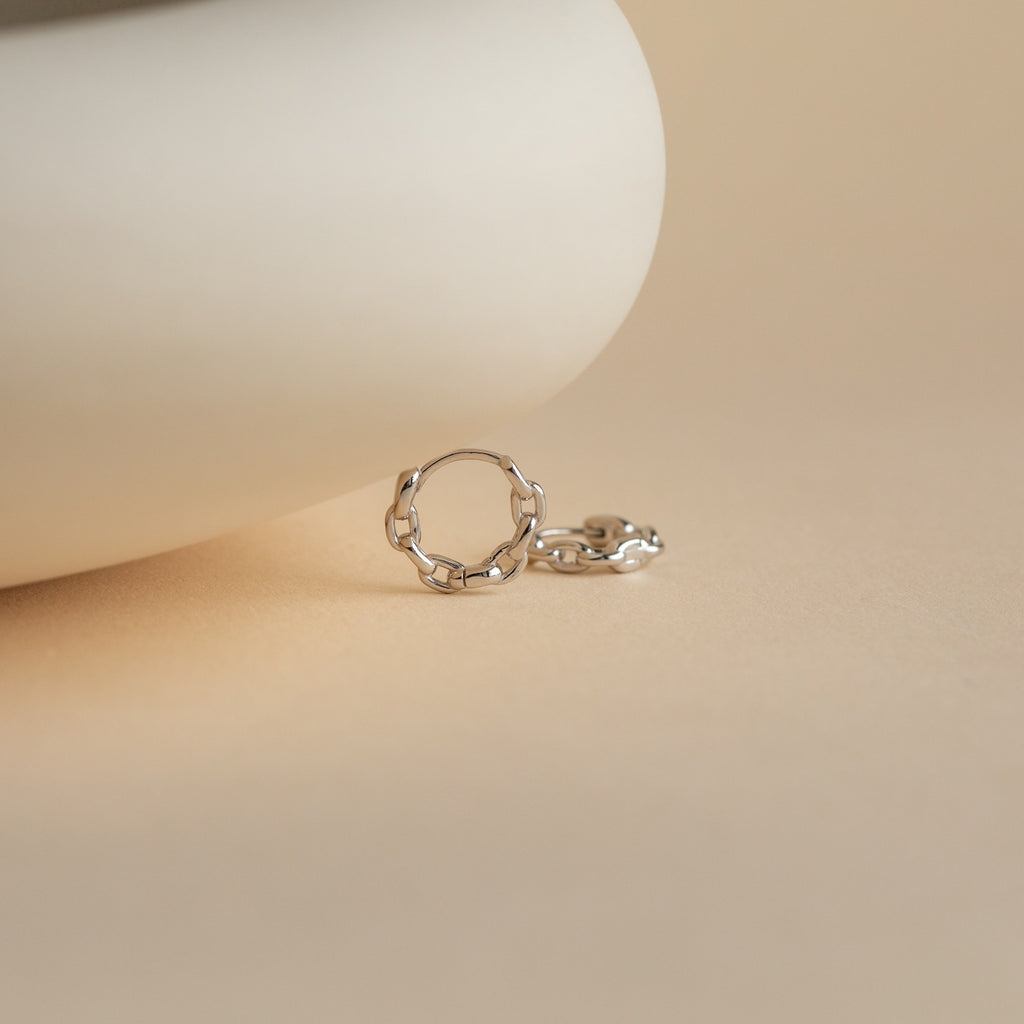 A single Link Huggies earring in silver rests on a smooth beige surface with a softly blurred background.