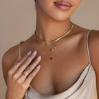 A woman wears the Little Red Heart Necklace layered with gold pieces and rings, styled with a satin dress and light pink nail polish.