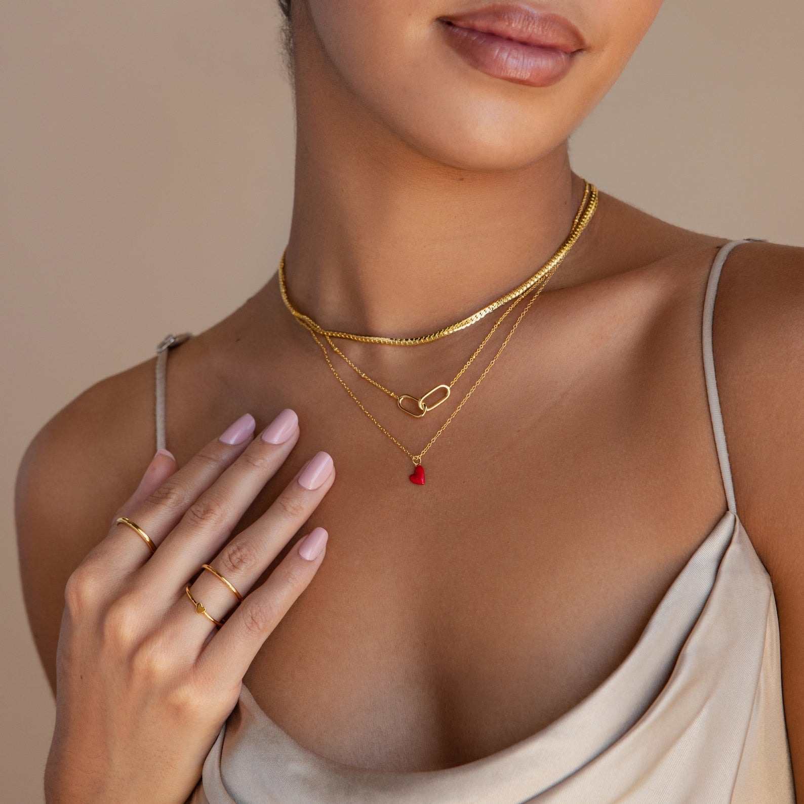 A woman wears the Little Red Heart Necklace layered with gold pieces and rings, styled with a satin dress and light pink nail polish.