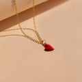 The Little Red Heart Necklace features a gold heart charm with a small red pendant, ideal for a minimalist look, shown on a beige surface.