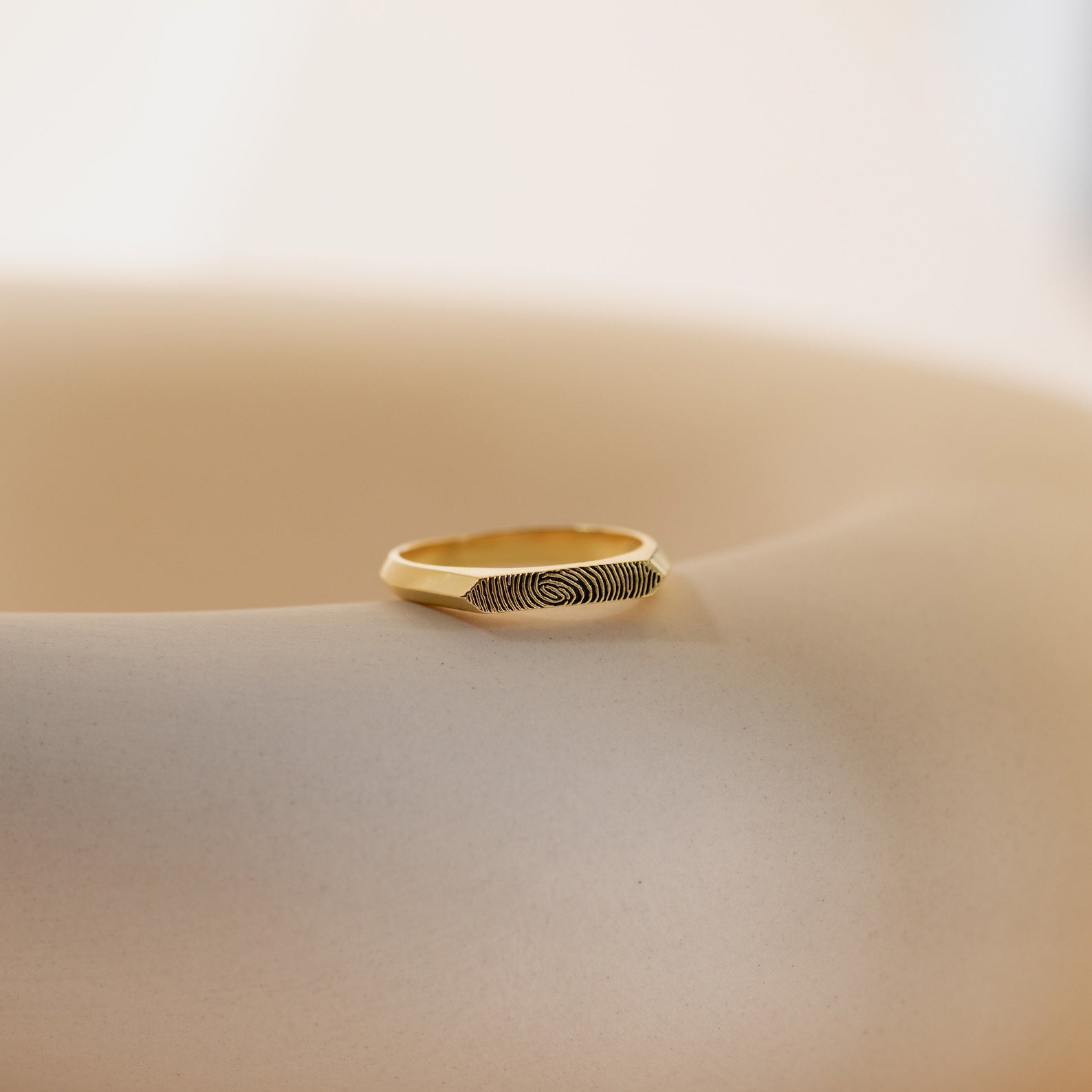 The Thin Fingerprint Signet Ring, engraved with your unique fingerprint, rests on a smooth beige surface in soft natural light—a personalized gift to treasure.
