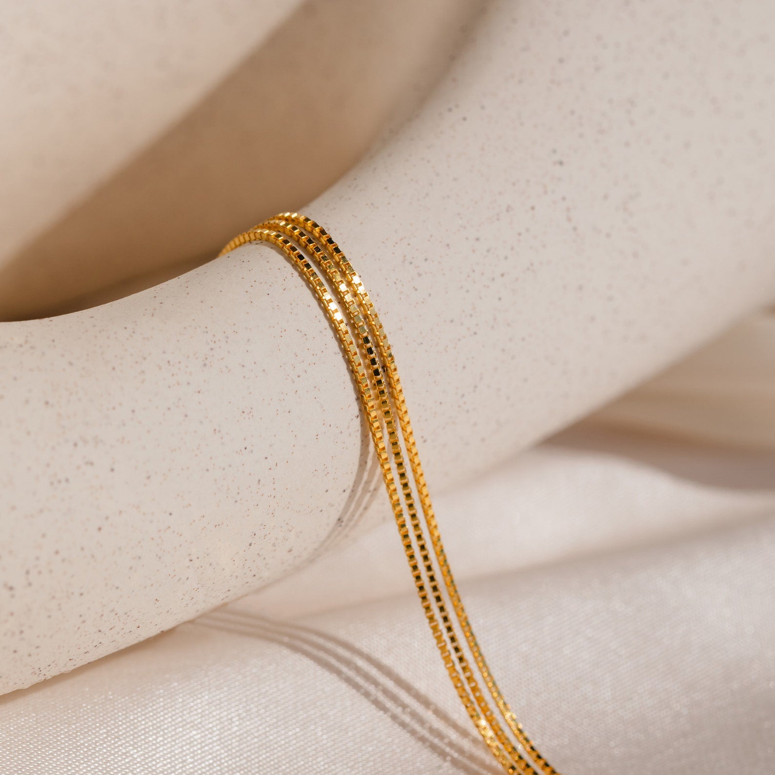 Close-up of the Triple Box Chain Necklace in gold, displayed on a smooth, light beige surface—ideal for layering with your favorite pieces.