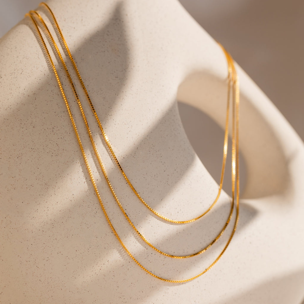 The Triple Box Chain Necklace, featuring three delicate gold layers, drapes elegantly over a smooth, abstract beige sculpture under soft lighting.