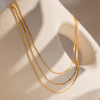 The Triple Box Chain Necklace, featuring three delicate gold layers, drapes elegantly over a smooth, abstract beige sculpture under soft lighting.